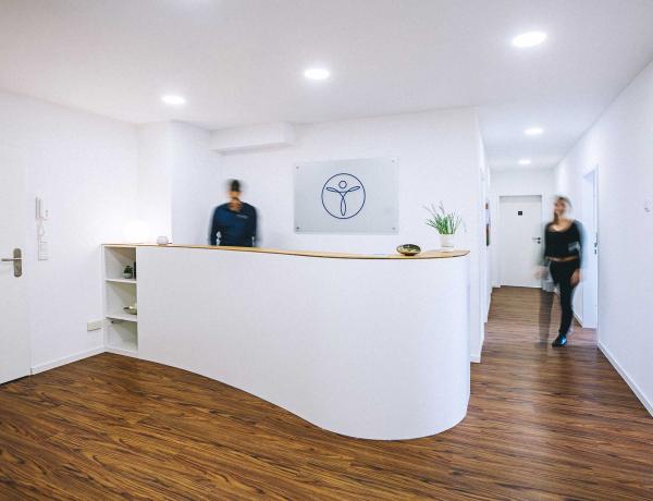 Links im Eck ist die Eingangstür, in der Mitte eine halbrunde Rezeption, darüber hängt das Logo der Praxis. Die Menschen sehen verschwommen aus.